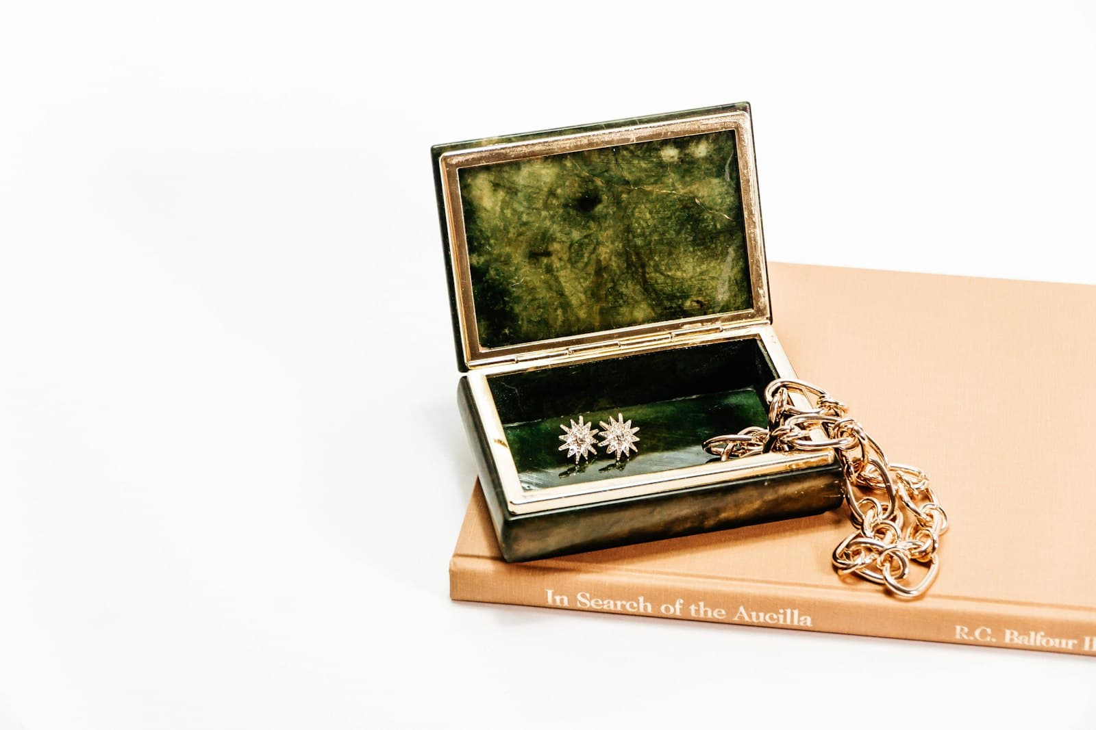 A small vintage jewelry box holding a pair of earrings and a gold chain, resting on an old book
