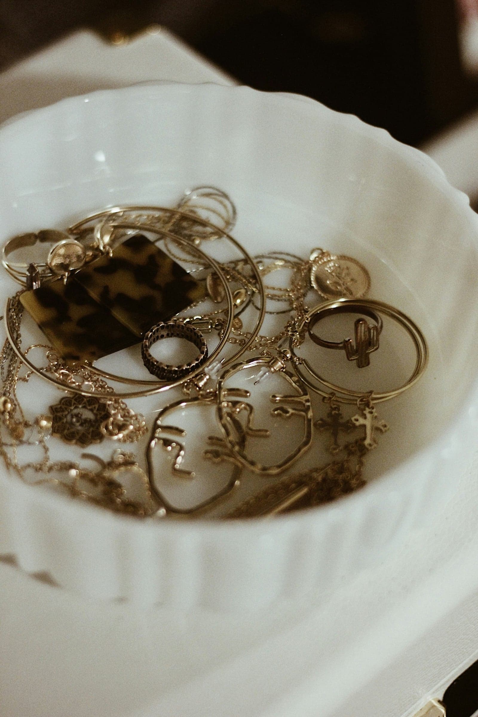 A white ceramic dish crowded with gold bangles, rings, chains, and sculptural face-shaped earrings