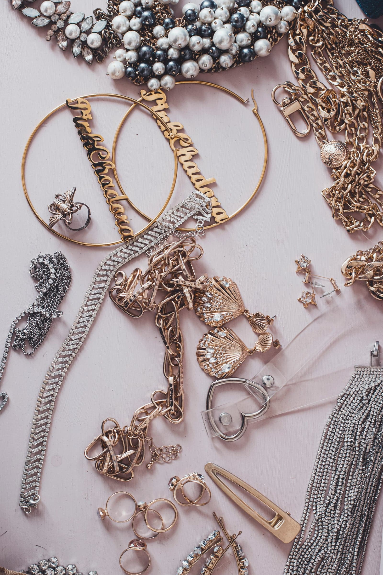 A maximalist flat lay of statement jewelry: oversized name hoops, rhinestone shell earrings, pearls, and tangled gold chains on a pink surface